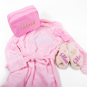 May include: Pink robe with a white "Mom" embroidered on the front, a pink cosmetic bag with "MOM" in rhinestones, a pink headband with "Mom" in glitter, and two white slippers with pink glitter "Mom" on the front.
