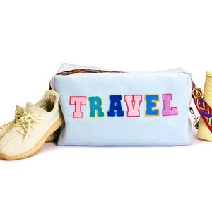 Puede incluir: Una bolsa de viaje azul claro con la palabra "TRAVEL" bordada en colores en la parte delantera. La bolsa tiene una correa colorida y est&aacute; junto a un par de zapatillas beige y un vaso amarillo con una pajita.