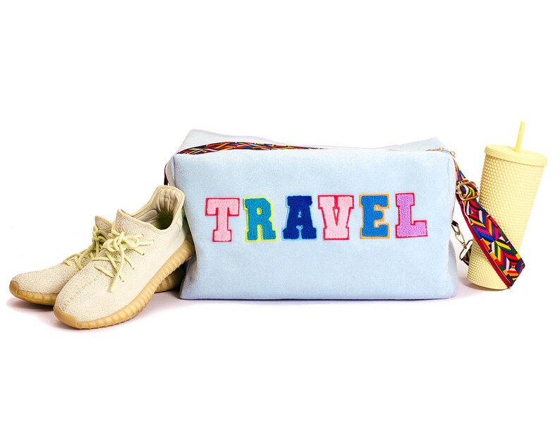 Puede incluir: Una bolsa de viaje azul claro con la palabra "TRAVEL" bordada en colores en la parte delantera. La bolsa tiene una correa colorida y est&aacute; junto a un par de zapatillas beige y un vaso amarillo con una pajita.