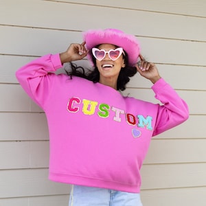 Peut inclure: Un sweat-shirt rose avec le mot "CUSTOM" en lettres colorées et un petit cœur violet. Le mannequin porte des lunettes de soleil en forme de cœur rose et un chapeau de cow-boy rose.