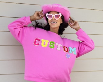 May include: A pink sweatshirt with the word "CUSTOM" in colorful letters and a small purple heart. The model is wearing a pink heart-shaped sunglasses and a pink cowboy hat.