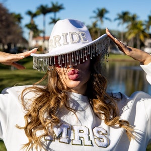 May include: A white cowboy hat with a rhinestone fringe and the word "BRIDE" spelled out in rhinestones. The person wearing the hat is also wearing a white sweatshirt with the word "MRS" spelled out in gold glitter.