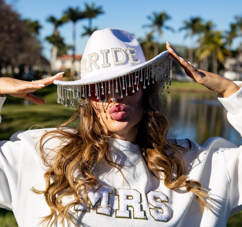 May include: A white cowboy hat with a rhinestone fringe and the word "BRIDE" spelled out in rhinestones. The person wearing the hat is also wearing a white sweatshirt with the word "MRS" spelled out in gold glitter.