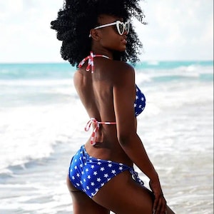 May include: A blue bikini with white stars and red and white striped ties. The model is wearing heart-shaped sunglasses. The background is a beach with blue water and a bright sky. The bikini is a patriotic theme.