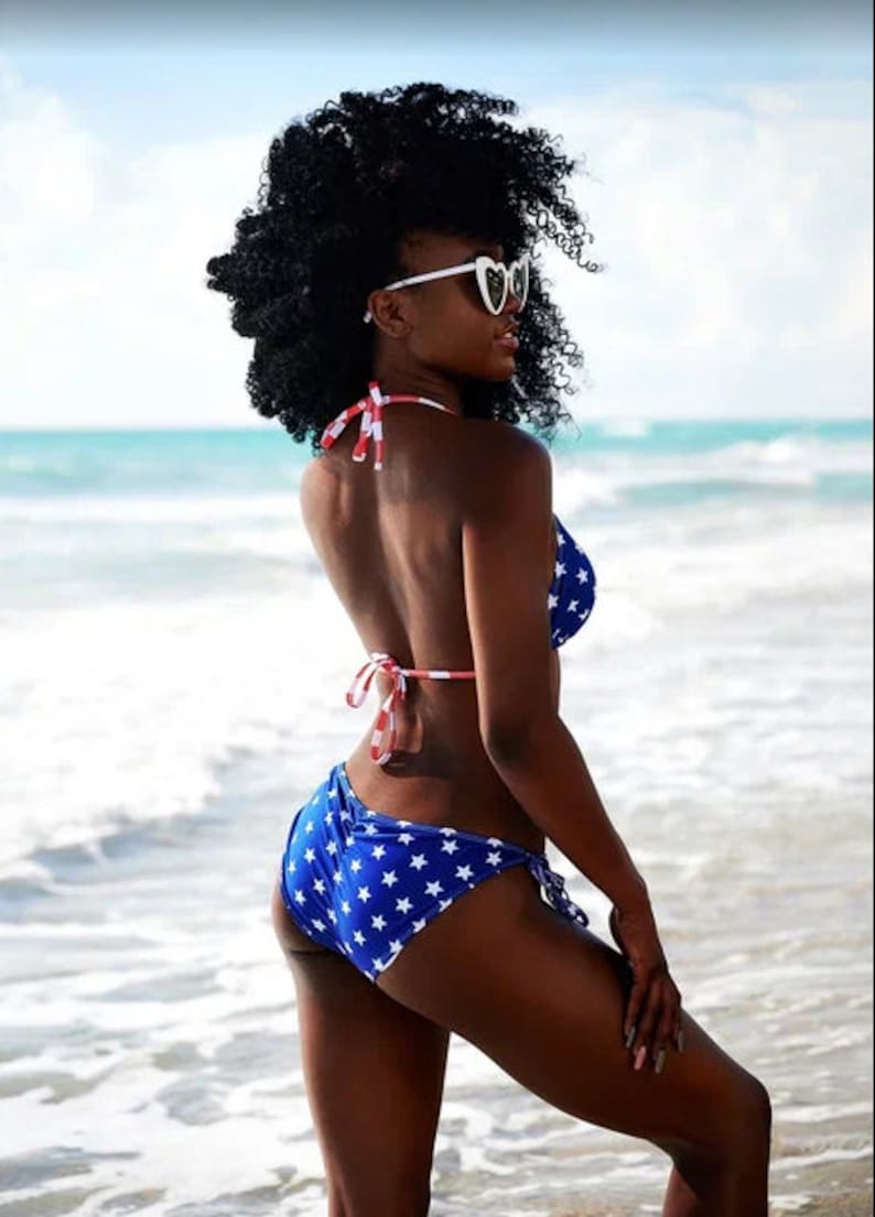 May include: A blue bikini with white stars and red and white striped ties. The model is wearing heart-shaped sunglasses. The background is a beach with blue water and a bright sky. The bikini is a patriotic theme.