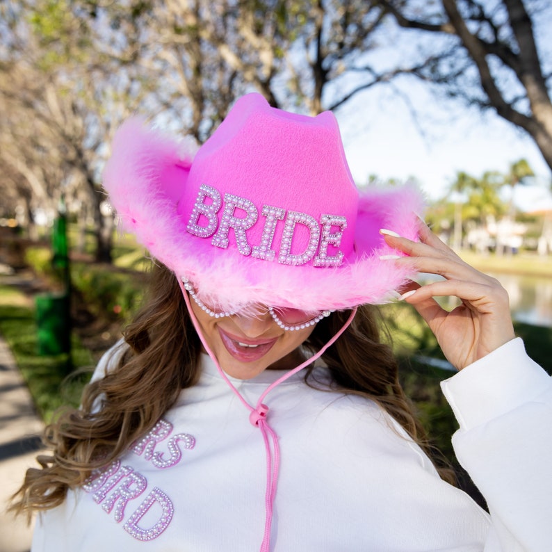 May include: A bright pink cowboy hat with the word "BRIDE" spelled out in sparkling rhinestones. The hat features a fluffy feather trim and a pink chin strap. The hat is part of a bridal theme.