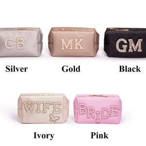 May include: Six cosmetic bags in different colors with jeweled lettering. The bags are silver, gold, black, ivory, and pink. The lettering on the bags reads "CB", "MK", "GM", "WIFE", and "BRIDE".
