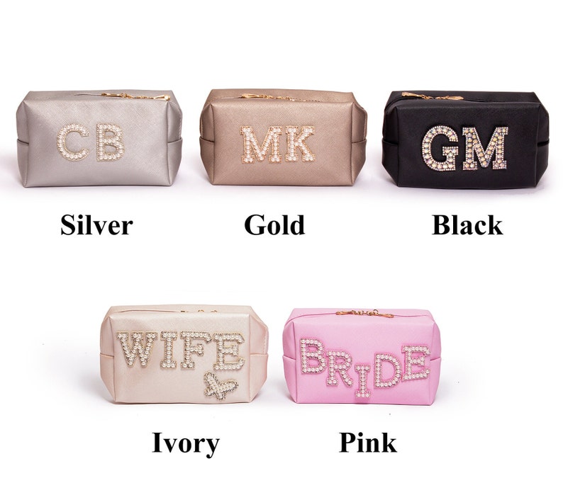 May include: Six cosmetic bags in different colors with jeweled lettering. The bags are silver, gold, black, ivory, and pink. The lettering on the bags reads "CB", "MK", "GM", "WIFE", and "BRIDE".