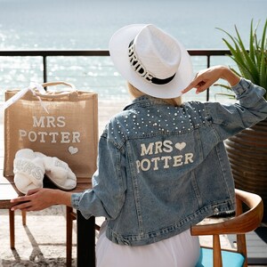 May include: A woman wearing a white hat with the word "Bride" on it, a denim jacket with pearl embellishments and the words "Mrs. Potter" on the back, and white slippers with the word "Mrs" on them. She is holding a burlap tote bag with the words "Mrs. Potter" on it.