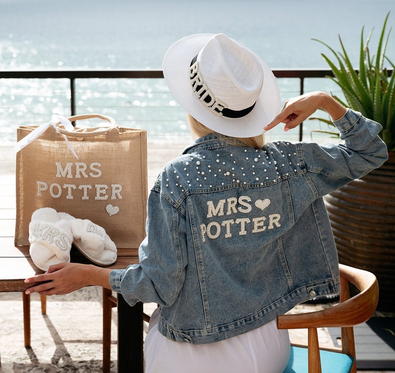 May include: A woman wearing a white hat with the word "Bride" on it, a denim jacket with pearl embellishments and the words "Mrs. Potter" on the back, and white slippers with the word "Mrs" on them. She is holding a burlap tote bag with the words "Mrs. Potter" on it.