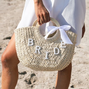 May include: A white straw tote bag with a wooden handle and a white satin ribbon. The bag has the word "BRIDE" spelled out in white letters.