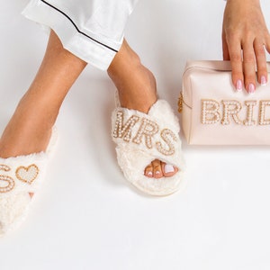 May include: A pair of white fuzzy slippers with gold and pearl embellishments. One slipper says "MRS" and the other says "S" with a heart. A pink satin clutch with gold and pearl embellishments says "BRIDE".