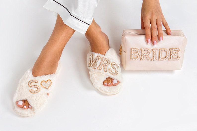 May include: A pair of white fuzzy slippers with gold and pearl embellishments. One slipper says "MRS" and the other says "S" with a heart. A pink satin clutch with gold and pearl embellishments says "BRIDE".