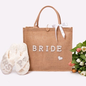 May include: A brown burlap tote bag with the word "BRIDE" in white letters and a small heart design. The bag is filled with white fluffy slippers with the words "MRS" and "BRIDE" on them. A bouquet of white and orange tulips is next to the bag.
