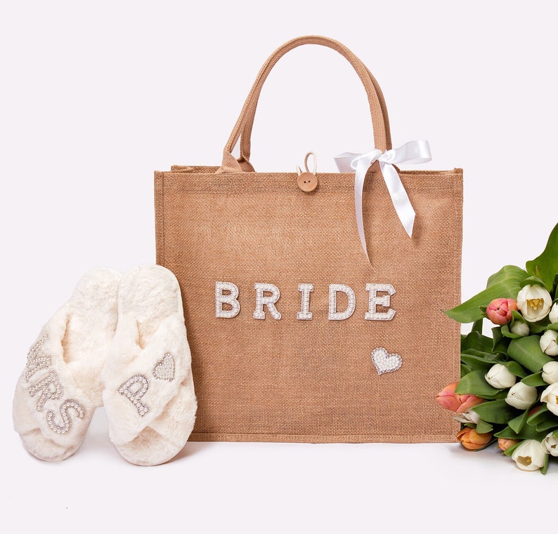 May include: A brown burlap tote bag with the word "BRIDE" in white letters and a small heart design. The bag is filled with white fluffy slippers with the words "MRS" and "BRIDE" on them. A bouquet of white and orange tulips is next to the bag.