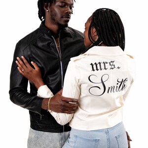 K&ouml;nnte beinhalten: Ein Paar posiert f&uuml;r ein Foto. Die Frau tr&auml;gt eine wei&szlig;e Lederjacke mit dem Text "mrs. Smith" auf dem R&uuml;cken. Der Mann tr&auml;gt eine schwarze Lederjacke.