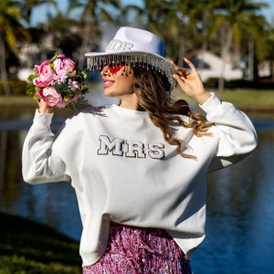 May include: A white sweatshirt with the word "MRS" in gold glitter letters. The model is wearing a white hat with a beaded trim and pink heart-shaped sunglasses. She is holding a bouquet of pink roses.