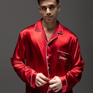 Puede incluir: Una camisa de pijama de manga larga con botones, confeccionada en tela de satén rojo vibrante con ribetes blancos en el cuello, los puños y la tapeta frontal. El bolsillo del pecho tiene bordado el nombre James en letras blancas.