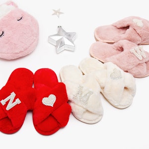 May include: Three pairs of fuzzy slippers with criss-cross straps. The slippers are in red, pink, and white. Each pair has a different letter or heart design with rhinestones.
