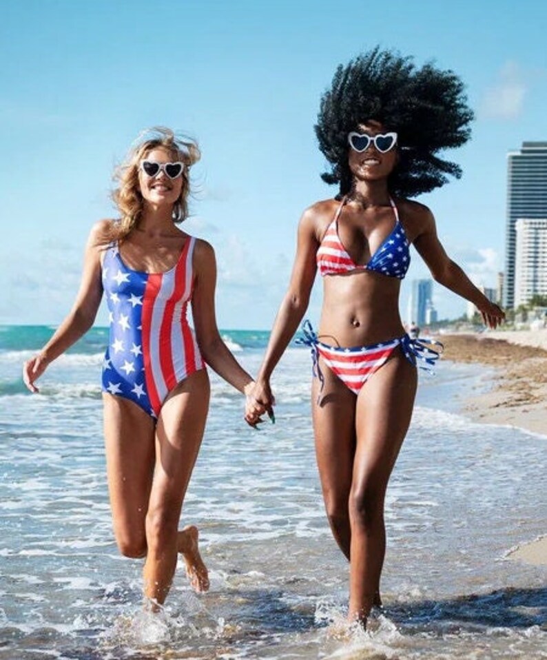 May include: Two people wearing patriotic swimwear run on a beach. One wears a one-piece swimsuit with a red, white, and blue American flag design. The other wears a bikini with a similar pattern. Both wear heart-shaped sunglasses.