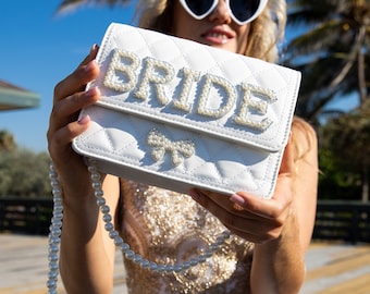 May include: White quilted handbag with a pearl embellished "BRIDE" design and a pearl bow. The bag has a pearl chain strap.