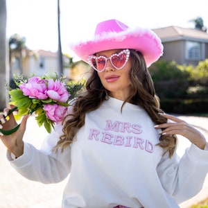 May include: A white sweatshirt with the text "MRS REBIRD" in pink rhinestones. The person is wearing a pink cowboy hat with white fur trim and heart-shaped sunglasses. A bouquet of pink peonies and greenery is held in the hand.