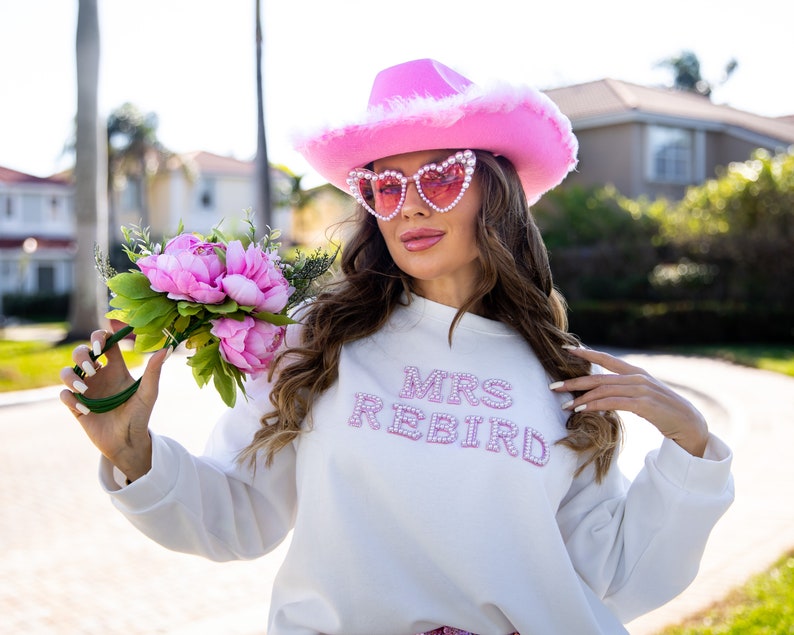 May include: A white sweatshirt with the text "MRS REBIRD" in pink rhinestones. The person is wearing a pink cowboy hat with white fur trim and heart-shaped sunglasses. A bouquet of pink peonies and greenery is held in the hand.