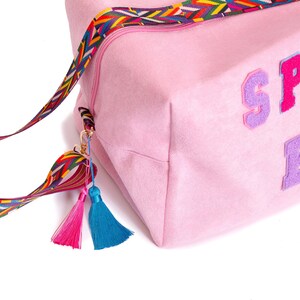 Puede incluir: Bolsa de deporte rosa con una correa y borlas de colores. La bolsa tiene la palabra "SPORT" escrita en letras de fieltro de colores.