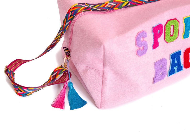 Puede incluir: Bolsa de deporte rosa con una correa y borlas de colores. La bolsa tiene la palabra "SPORT" escrita en letras de fieltro de colores.