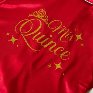 Puede incluir: Bata de sat&eacute;n roja con texto dorado brillante y detalles de estrellas. El texto dice "Mis Quince" con una corona encima de "Quince". Ribetes blancos bordean la bata.