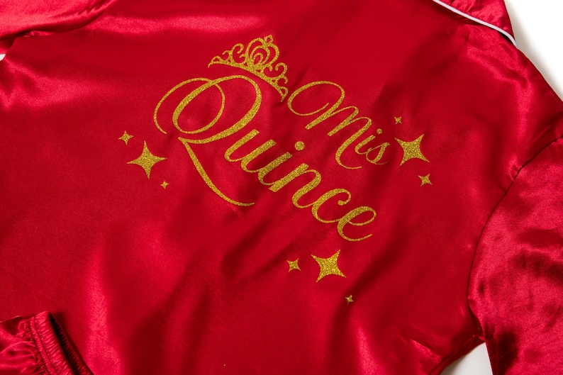 Puede incluir: Bata de sat&eacute;n roja con texto dorado brillante y detalles de estrellas. El texto dice "Mis Quince" con una corona encima de "Quince". Ribetes blancos bordean la bata.