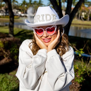 May include: A white cowboy hat with the word "BRIDE" spelled out in rhinestones and pearls. The hat is worn by a person wearing pink heart-shaped sunglasses with pearl accents.