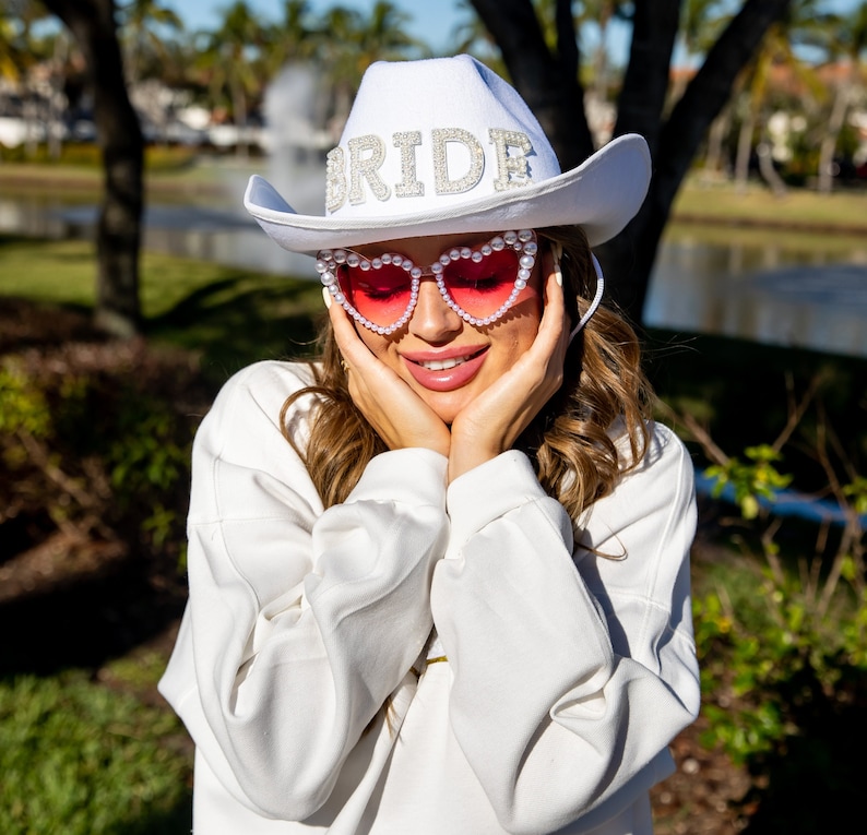May include: A white cowboy hat with the word "BRIDE" spelled out in rhinestones and pearls. The hat is worn by a person wearing pink heart-shaped sunglasses with pearl accents.