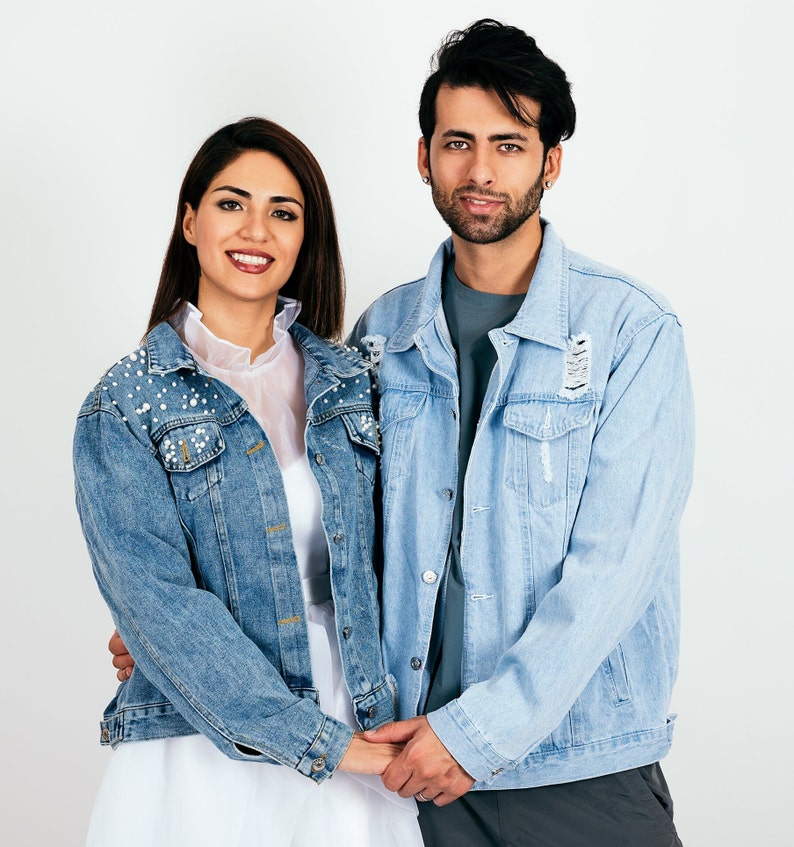 K&ouml;nnte beinhalten: Ein Paar tr&auml;gt blaue Jeansjacken, die Jacke der Frau ist mit Perlen verziert, sie stehen eng beieinander und l&auml;cheln in die Kamera.