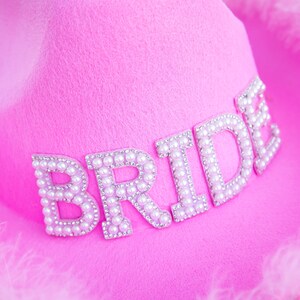 May include: A bright pink cowboy hat with the word "BRIDE" spelled out in pearl-like beads. The hat is trimmed with fluffy pink feathers, perfect for a bachelorette party or bridal shower.