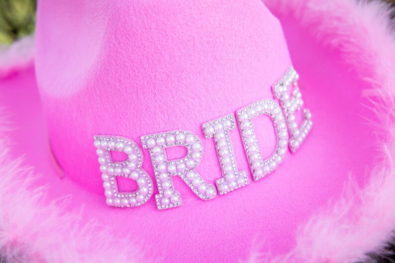 May include: A bright pink cowboy hat with the word "BRIDE" spelled out in pearl-like beads. The hat is trimmed with fluffy pink feathers, perfect for a bachelorette party or bridal shower.