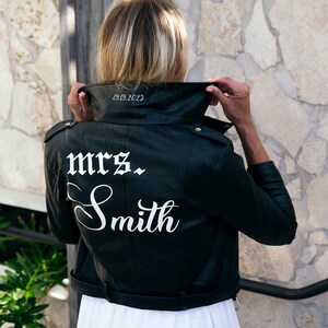 K&ouml;nnte beinhalten: Schwarze Lederjacke mit einer wei&szlig;en Aufschrift auf der R&uuml;ckseite, die "mrs. Smith" lautet, und dem Datum "05.09.2023" am Kragen.