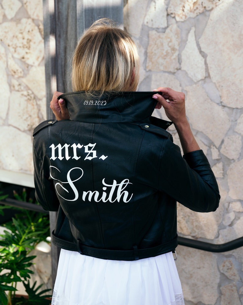 K&ouml;nnte beinhalten: Schwarze Lederjacke mit einer wei&szlig;en Aufschrift auf der R&uuml;ckseite, die "mrs. Smith" lautet, und dem Datum "05.09.2023" am Kragen.