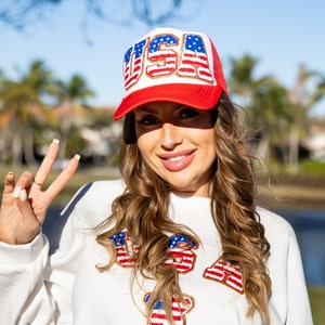May include: A woman wearing a white sweatshirt with a red, white, and blue "USA" graphic. She is wearing a red, white, and blue trucker hat with the word "USA" on it. She is giving a peace sign with her right hand.