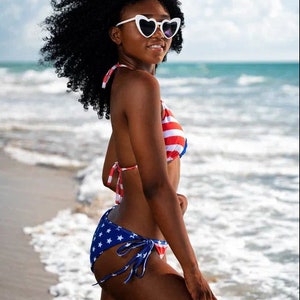 May include: A woman wearing a patriotic-themed bikini stands on a beach. The top is red and white striped, while the bottoms are blue with white stars. She wears heart-shaped sunglasses.