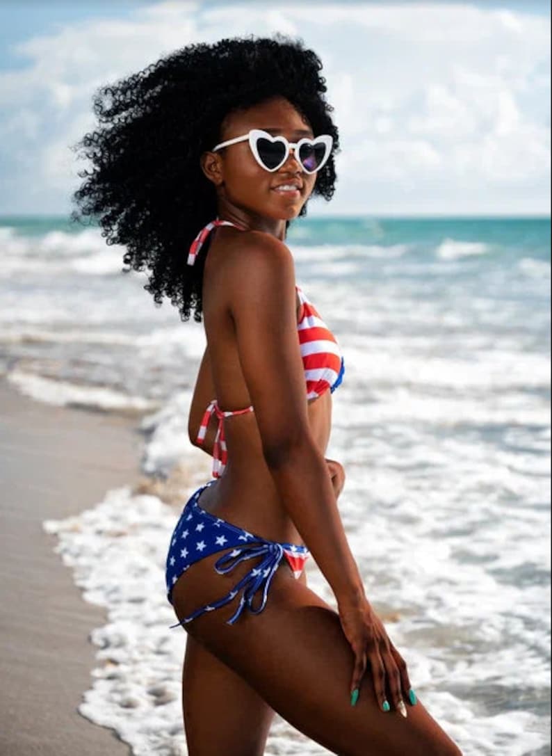 May include: A woman wearing a patriotic-themed bikini stands on a beach. The top is red and white striped, while the bottoms are blue with white stars. She wears heart-shaped sunglasses.