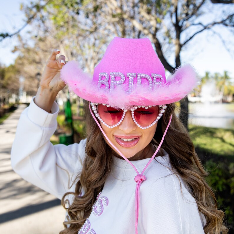May include: A pink cowboy hat with the word "BRIDE" spelled out in silver rhinestones. The hat has a fluffy pink feather trim and is paired with pink heart-shaped sunglasses with pearl accents. The sunglasses have a pink string.