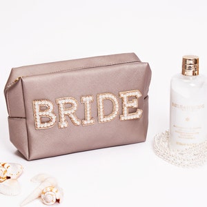 May include: A light brown cosmetic bag with the word "BRIDE" spelled out in gold letters with pearl accents. The bag is sitting on a white surface next to a bottle of body lotion and a pearl necklace.