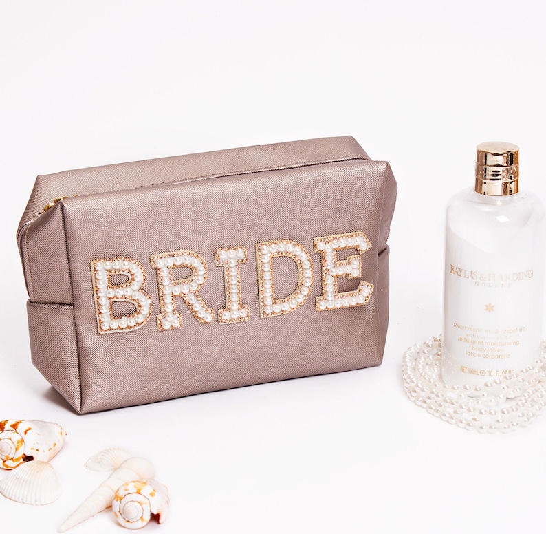 May include: A light brown cosmetic bag with the word "BRIDE" spelled out in gold letters with pearl accents. The bag is sitting on a white surface next to a bottle of body lotion and a pearl necklace.