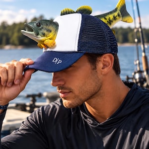 May include: A navy blue and white trucker hat featuring a three-dimensional largemouth bass fish attached to the front panel. The navy blue brim displays the white text D|P. The hat has a white front panel and a navy blue mesh back.