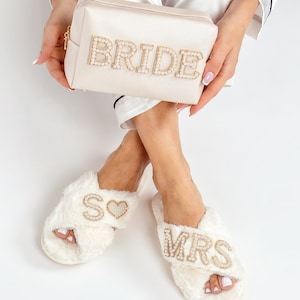 May include: White faux fur slippers with gold and pearl embellishments. One slipper says "MRS" and the other says "SO" with a heart. A white satin cosmetic bag with gold and pearl embellishments says "BRIDE".