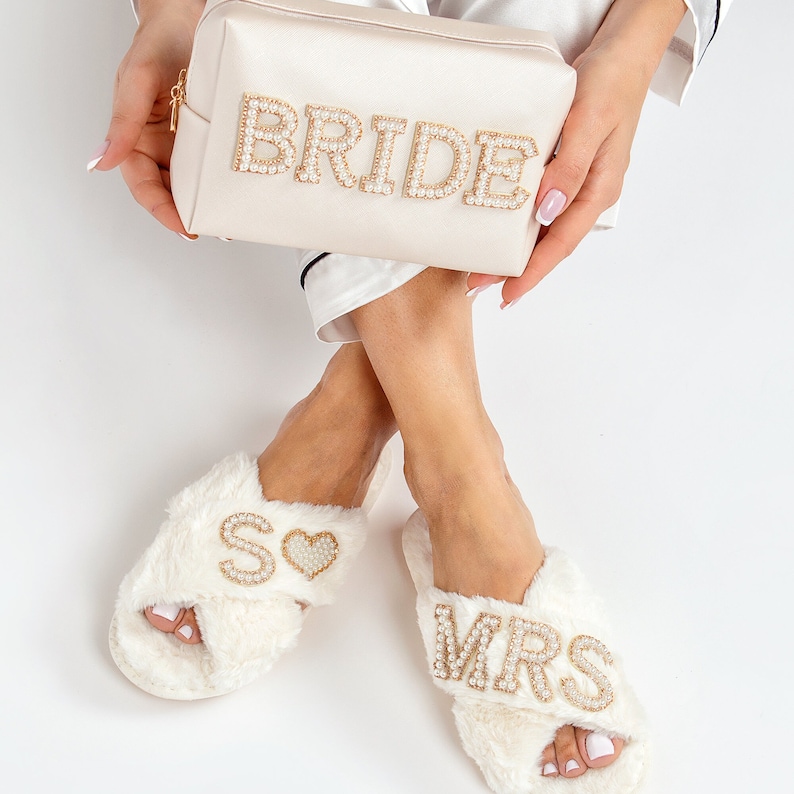 May include: White faux fur slippers with gold and pearl embellishments. One slipper says "MRS" and the other says "SO" with a heart. A white satin cosmetic bag with gold and pearl embellishments says "BRIDE".
