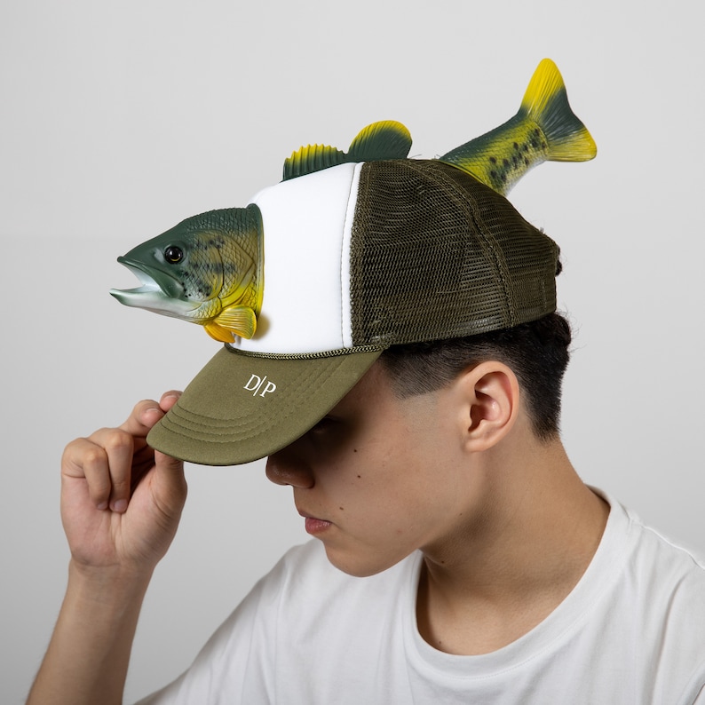 May include: A green and white trucker hat with a fish attached to the top. The fish is green, yellow, and brown. The hat has a white mesh back and a green front with the letters "D|P" embroidered on the front.
