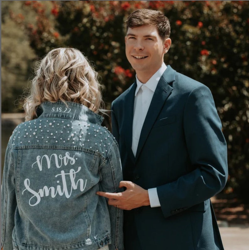 K&ouml;nnte beinhalten: Eine Braut tr&auml;gt eine Jeansjacke mit Perlenbesatz und dem Text "Mrs. Smith" sowie dem Datum "8.18.22" auf der R&uuml;ckseite. Die Braut steht neben einem Br&auml;utigam in einem dunkelblauen Anzug.