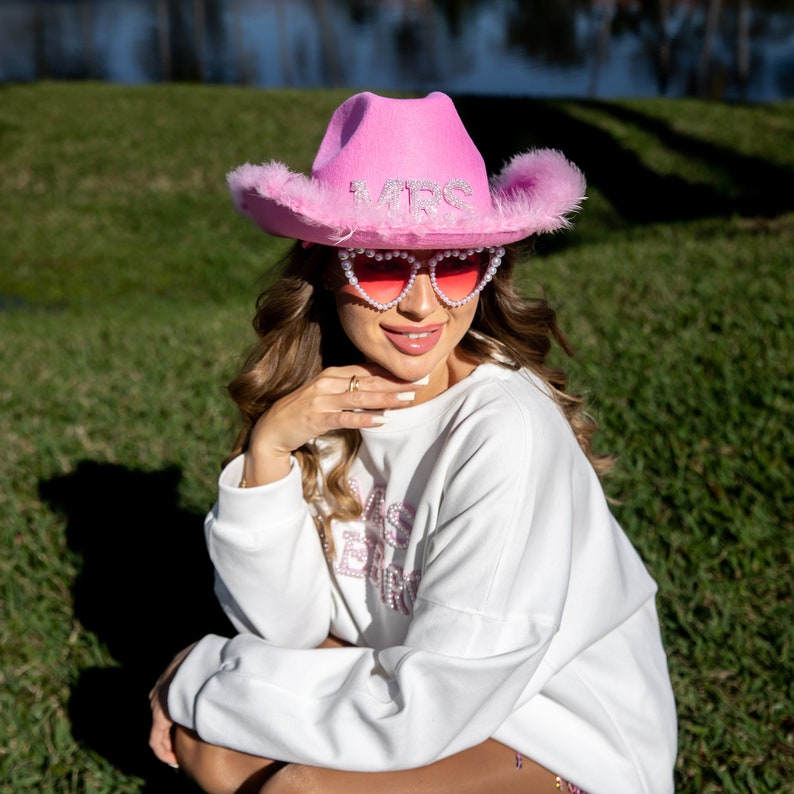 May include: A pink cowboy hat with the word "MRS" in rhinestones. The hat has a fluffy feather trim and is paired with heart-shaped sunglasses with pearl accents. A white sweatshirt with the text "Mrs. Bride" in rhinestones is also visible.